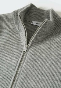 Suéter gris con cremallera, con textura acanalada, cuello alzado y cremallera metálica. La etiqueta muestra "MNG" en el interior.