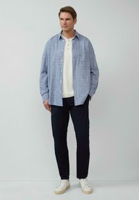 Hellblause, strukturierte Langarmbluse über einem weißen Henley, kombiniert mit dunklen Hosen und weißen Sneakern. Enthält eine Brusttasche.