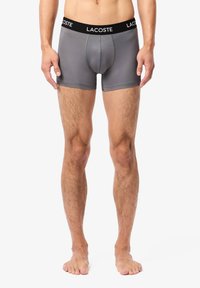 Szürke férfi boxer alsónadrág fekete "LACOSTE" márkajelzéssel ellátott derékkal, sima anyagból készült; testhezálló fazon rövid szárakkal és kontúros zsebbel.