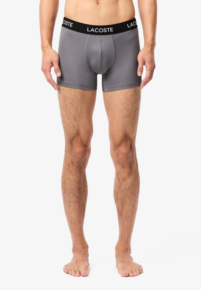 Boxers gris pour hommes avec une ceinture noire à logo "LACOSTE", en tissu lisse ; coupe ajustée avec des jambes courtes et une poche contournée.