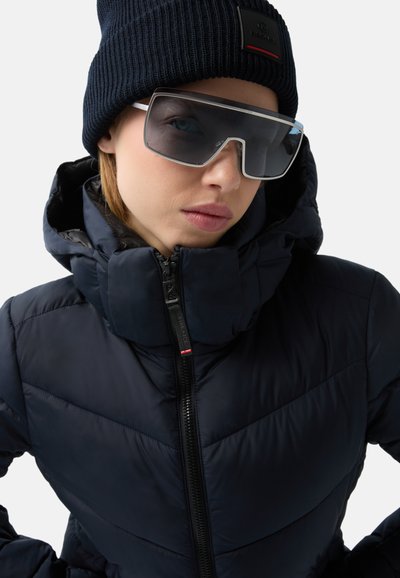Marineblaues Puffer-Jacke mit hohem Kragen, ausgestattet mit einem Reißverschluss und einem markanten, gesteppten Muster. Kombiniert mit einer schwarzen Strickmütze und übergroßen Sonnenbrillen.