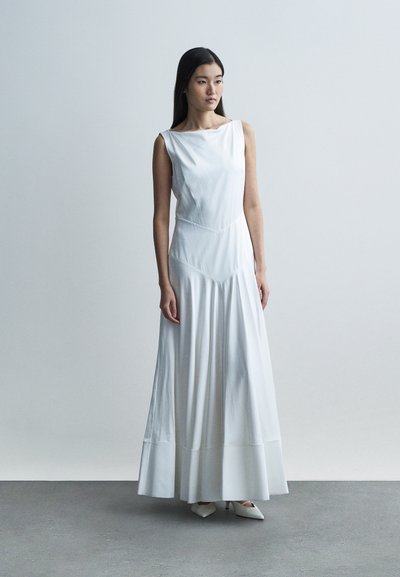 Vestido branco sem mangas com decote canoa, saia fluida e costuras geométricas. O tecido aparenta ser suave e leve. Sapatos brancos pontiagudos.