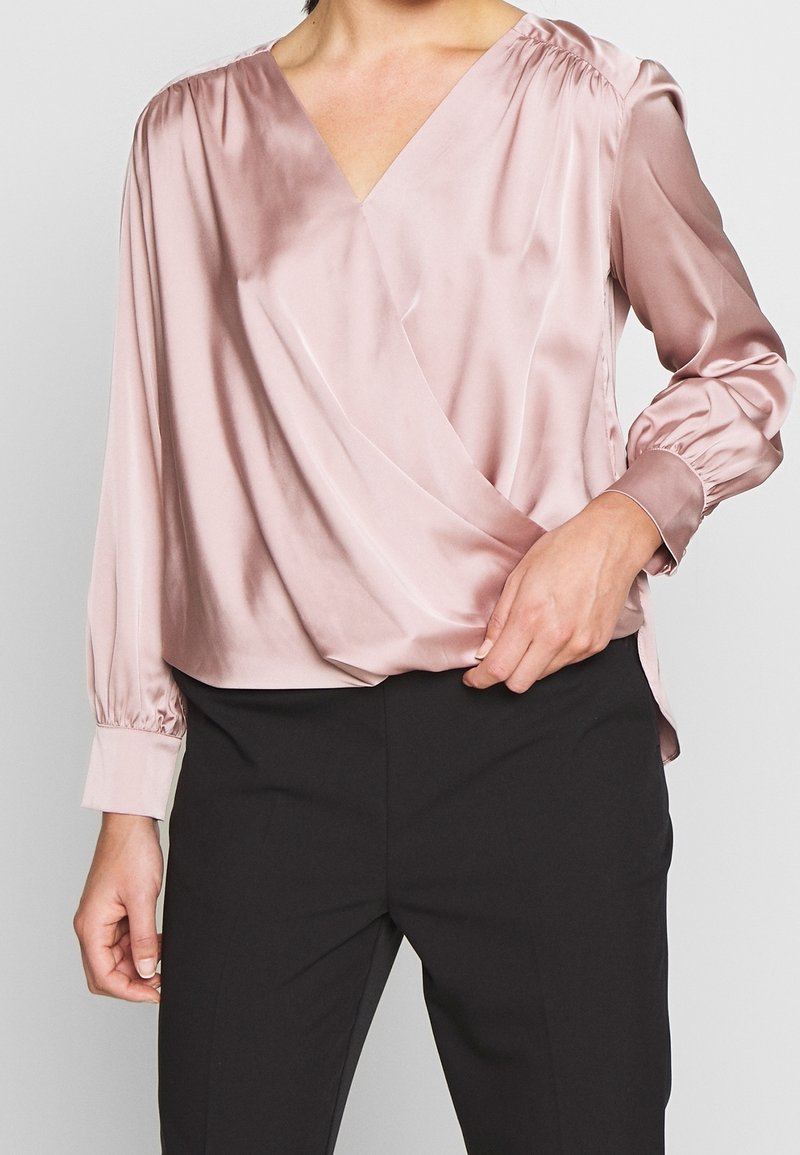Blouse en satin rose clair avec un col en V, des manches longues bouffantes et des épaules plissées ; se marie avec un pantalon noir ajusté pour un look soigné.