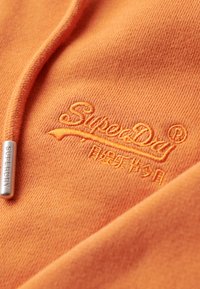 Orange hoodie med tekstureret stof; har et broderet "SuperDry" logo og et metallisk mærke med brandnavnet.