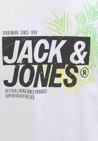 Valkoinen t-paita, jossa on musta ja keltainen "JACK & JONES" -teksti sekä vihreitä palmulehden grafiikoita. Sisältää tavaramerkin ja lisäbrändäystekstejä.