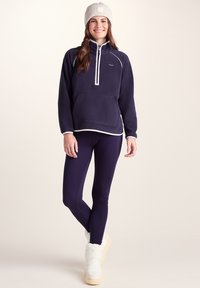 Woman smiling wearing a gray knit beanie, navy blue fleece pullover, dark leggings, and white winter boots against a plain light background.