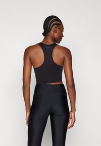 Sujeción deportiva negra con un diseño de espalda nadadora y un ajuste recortado. La textura parece suave, con un sutil detalle del logo en la parte posterior. Leggings negros visibles.