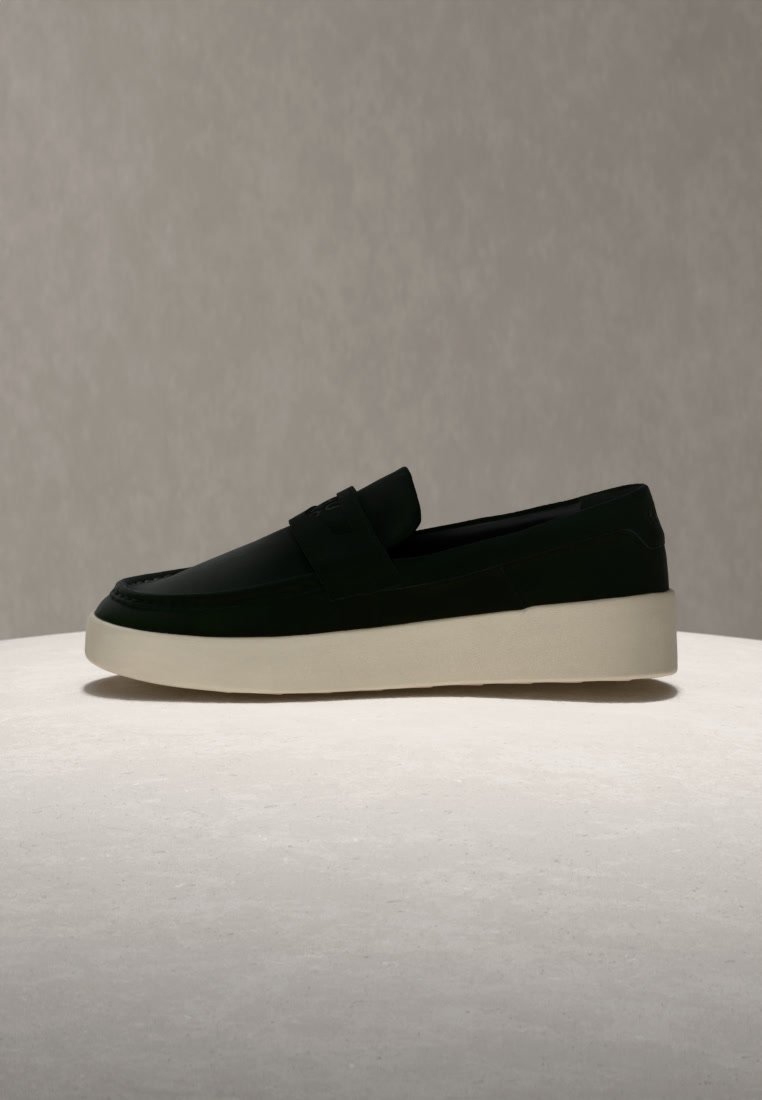 Mocassin noir sans lacets avec une épaisse semelle blanche présenté sur un fond gris uni.