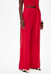 Red wide-leg trousers made of smooth fabric, featuring a high waist and a knotted tie detail at the front. Pairs with patterned shoes.