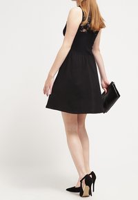 Schwarzes Kleid mit tailliertem Oberteil und ausgestelltem Rock, mit Spitzenakzenten im Rücken. Getragen mit schwarzen Stilettos und einer Clutch.