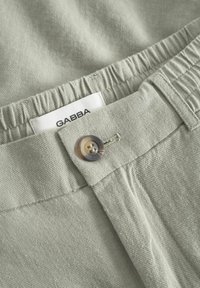 Pantalon avec taille élastique gris clair, fermeture par bouton et étiquette blanche affichant le nom de la marque "GABBA".