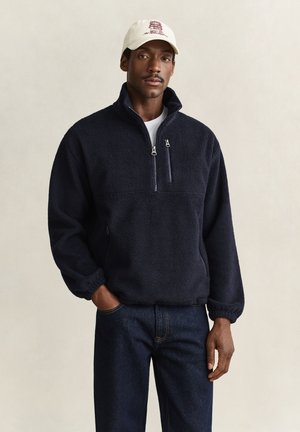 Homme portant un pull polaire bleu marine avec des poches zippées, un jean foncé et une casquette de baseball blanche, debout devant un fond uni.