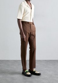 Kortärmad, krämfärgad texturerad skjorta, bruna skräddarsydda byxor och svarta loafers med färgglada accenter och dekorativ hårdvara.