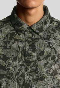 Närbild av en person som bär en svart skjorta med krage med ett geometriskt mönster i grönt och vitt samt tre svarta knappar.