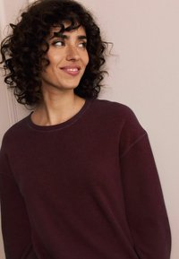 Burgundi bordázott pulóver széles nyakkivágással és laza fazonnal. Göndör haj és egy enyhe mosoly egészíti ki az egyszerű dizájnt.