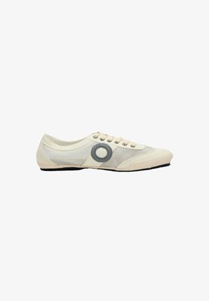 Zapatilla de corte bajo de color beige claro con paneles de malla, cordones beige y un logo circular oscuro en el lateral, sobre un fondo blanco.