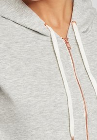 Grijze zip-up hoodie met een gestructureerde stof, voorzien van een roségouden rits en witte koorden. Close-up van de halslijn en het ritsgebied.