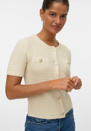 Cardigan crème à manches courtes avec un tricot texturé, col rond, boutons dorés et deux poches avant, associé à un denim.