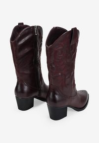 Botas de vaquero de cuero burdeos con tacón medio, costuras decorativas, cremallera lateral y punta redondeada; textura suave con patrones bordados.