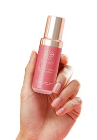 Bottiglia di siero rosa con un tappo dorato, caratterizzata da un logo floreale e testo bianco. Tenuta in mano con unghie naturali su uno sfondo semplice.