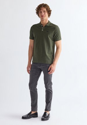 Mann steht und trägt ein olivgrünes kurzärmeliges Poloshirt, graue Slim-Fit-Jeans und schwarze Leder-Loafers vor weißem Hintergrund.