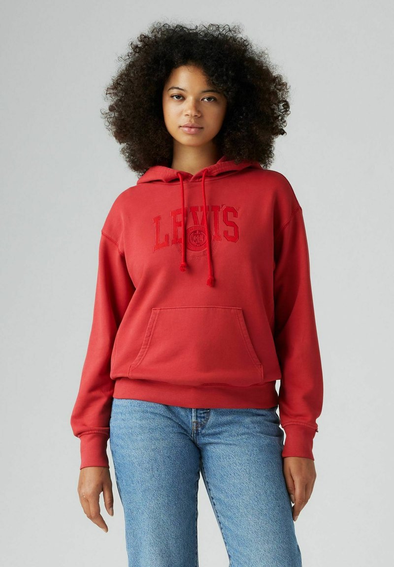 Felpa rossa in cotone con cappuccio, con coulisse e tasca frontale, caratterizzata dal logo "LEVIS" in testo rosso abbinato. Abbinata a jeans in denim azzurri chiari.