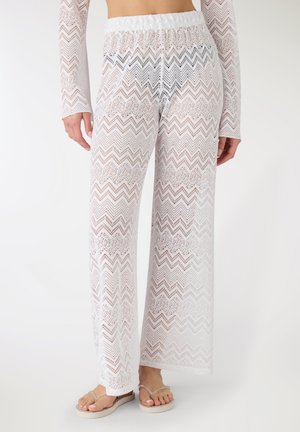 Donna che indossa pantaloni bianchi trasparenti in crochet con motivo a zigzag e top abbinato a maniche lunghe, abbinati a infradito beige, in piedi davanti a uno sfondo bianco.