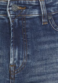 Gros plan sur un jean en denim bleu avec un bouton métallique visible, une boucle de ceinture, des coutures jaunes et des détails de poche avant.