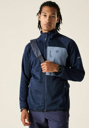 Junger Mann trägt eine marineblaue Zip-Jacke mit einer hellblauen Brusttasche und einem dunklen Schultertaschenriemen quer über der Brust.