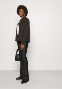 Καφέ υφασμάτινο μπουφάν πάνω από λευκό μπλουζάκι, συνδυασμένο με μαύρα φαρδιά παντελόνια και μποτάκια μέχρι τον αστράγαλο. Κρατάει μια μαύρη τσάντα με το ένα χέρι.