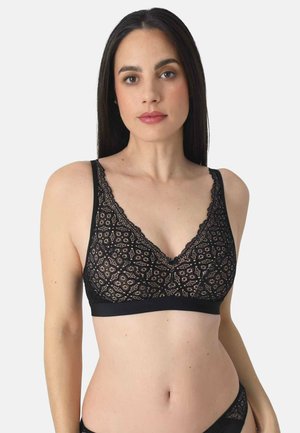 Frau mit langen dunklen Haaren, die ein schwarzes Spitzen-Bralette und passende Unterwäsche vor einem einfarbigen hellen Hintergrund trägt.