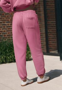 Ružové teplákové nohavice vyrobené z mäkkého materiálu s úzkym strihom, elastickým pásom a manžetami na členkoch; obsahujú zadnú kapsu a sú skombinované s bielymi teniskami.