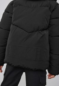 Zwarte donsjas met doorgestikt ontwerp, voorzien van een opvallend reliëf met de tekst "BAKER" bij de zoom en een gladde textuur. Inclusief verstelbare capuchon.