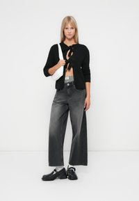 Cardigan preto em malha com fecho de amarração, jeans cinza de perna larga, sapatos pretos e bolsa branca. O fundo simples realça os detalhes do conjunto.