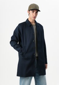 Casaco azul marinho, até ao joelho, com botões e gola. O material parece suave, com bolsos frontais e um corte descontraído.