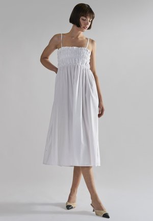 Kvinde iført hvid ærmeløs midi-kjole med smocket bodice, stående med den ene hånd bag ryggen og beige hæle med sorte tåhætte.