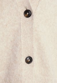 Cardigan en laine beige clair présentant un décolleté en V et trois boutons noirs sur le devant. Texture douce avec un motif tricoté.