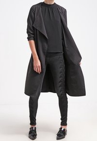 Dlouhý černý kabát s přehozeným límcem, nošený přes černou blůzu a těsné kalhoty s texturovanými detaily, doplněný lesklými černými botami.