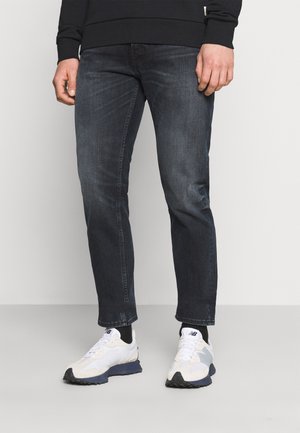 Jeans de ganga azul escuro com um corte slim e um leve desbotado, combinados com ténis brancos e cinzentos com uma sola texturada e detalhes em contraste.