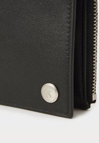 Black leather wallet featuring a silver logo button and a zippered closure. The texture is smooth with minimal stitching details.