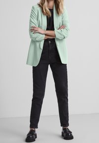 Světlý mátový blazer s otevřeným předním dílem a ohrnutými rukávy, spárovaný s černými džíny se zvýšeným pasem a robustními černými botami. Jednoduchý a moderní design.