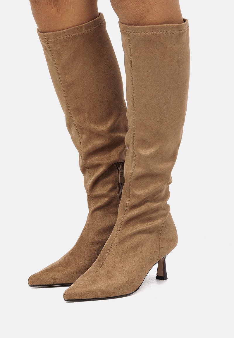 Botas de ante en color marrón claro, de caña alta hasta la rodilla, con puntera afilada y tacones delgados, llevadas por una persona sobre un fondo blanco liso.