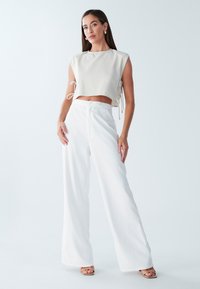 Creme crop top med sidesnore, parret med hvide wide-leg bukser med en tekstureret finish. Modellen står selvsikkert foran en lys baggrund.
