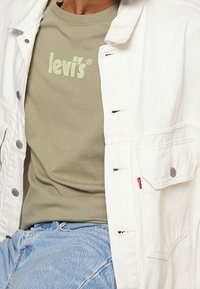 Beige tričko Levi's nošené pod rozepnutou bílou džínovou bundou a kombinované se světle modrými džíny.