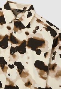 Camicia con bottoni in un tessuto leggero, con un design a stampa di mucca in tonalità marroni e beige. Bottoni neri e colletto standard.