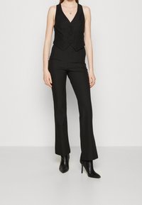 Svart skräddarsydd väst med V-ringning, knappstängning och utsvängda byxor. Slät tyg med en elegant design. Bärs med spetsiga svarta stövlar.