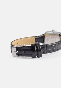Correa de reloj de cuero negro con acabado texturizado, hebilla de metal plateado y forro de cuero marrón. La forma es rectangular con una superficie lisa.