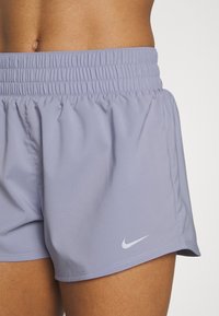 Ljuslila idrottsshorts med elastisk midja, med en liten vit Nike-logga på nedre delen av högra benet. Slät textur, avslappnad design.