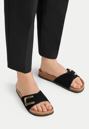 Black slide sandals with a suede upper, featuring a gold metal buckle and a cork sole. Accompanied by black tailored pants.