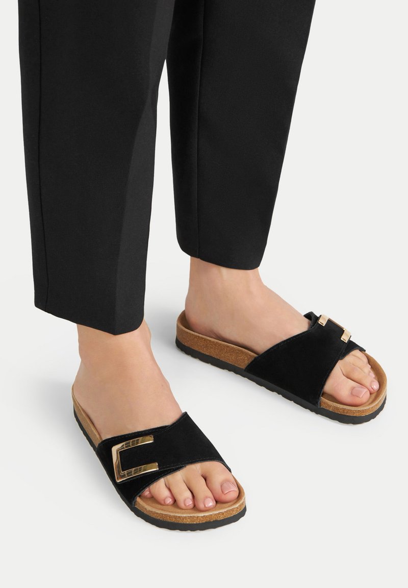 Black slide sandals with a suede upper, featuring a gold metal buckle and a cork sole. Accompanied by black tailored pants.